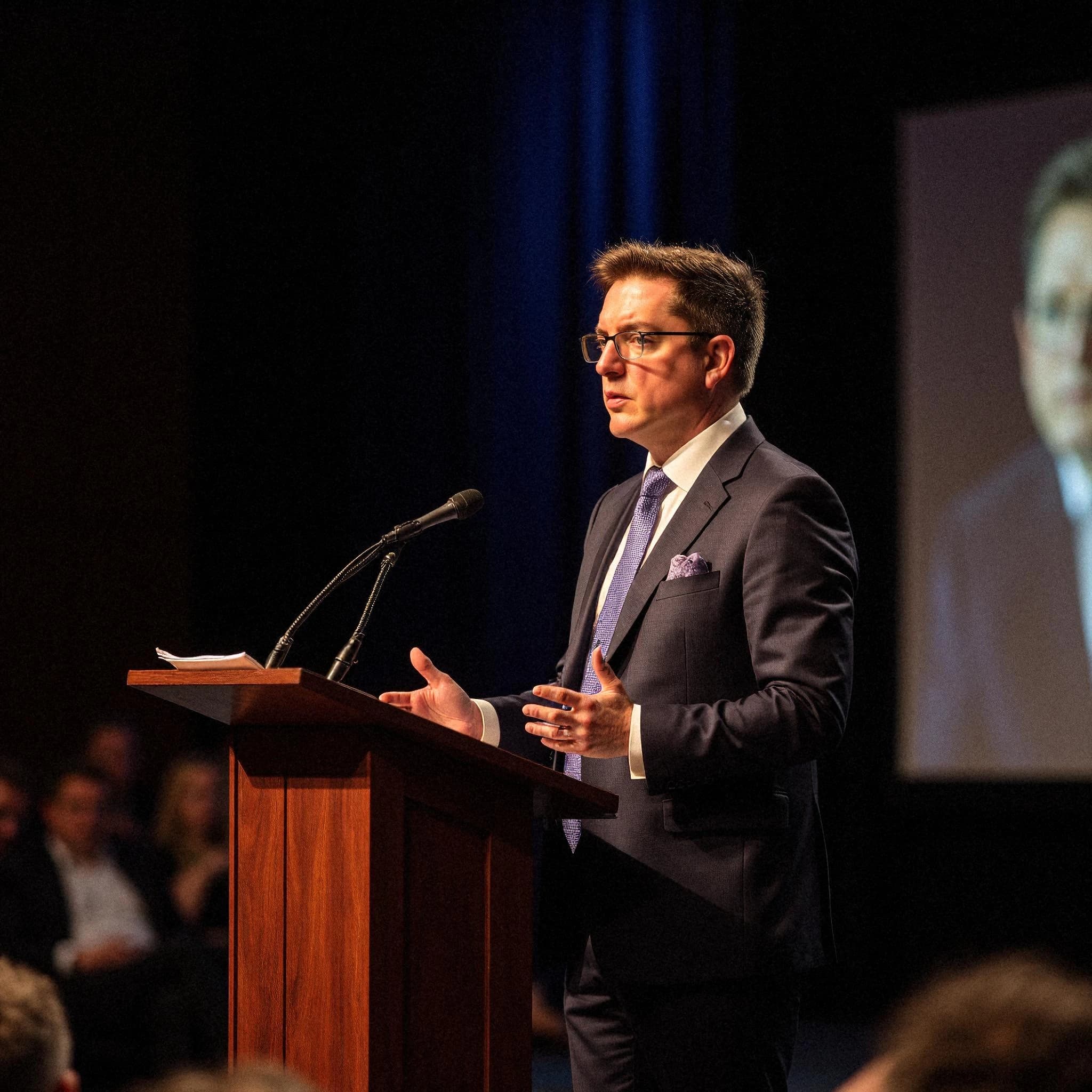 Professional headshot example: Man Stage Suit Speaking Podium — LinkedInHeadshots.ai